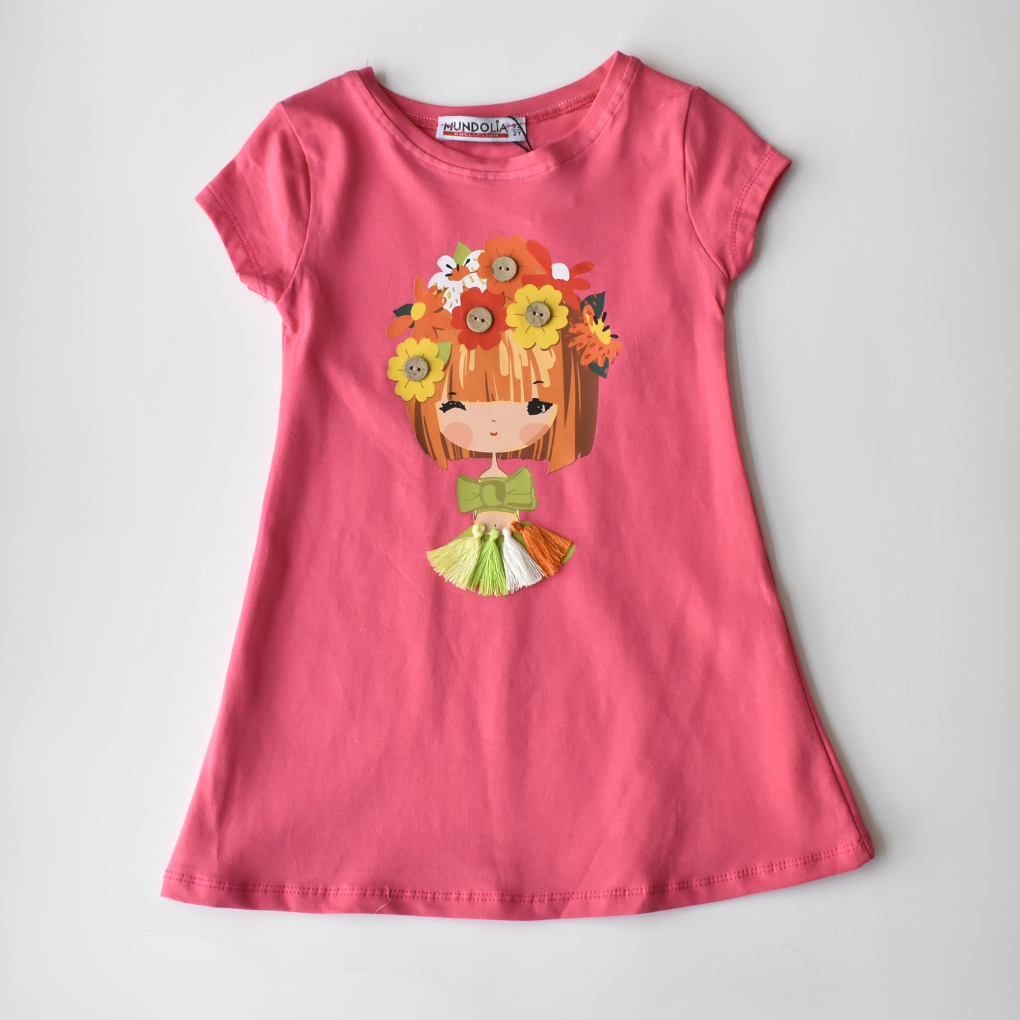 Pink short-sleeve girls’ dress with a cartoon girl graphic wearing a floral headpiece, green bow, and multicolor tassel skirt detail.