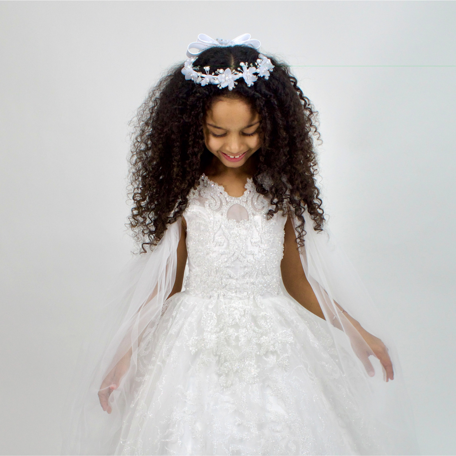 Young girl in a white lace communion dress with tulle details, wearing a floral headband with bow, smiling while looking down.