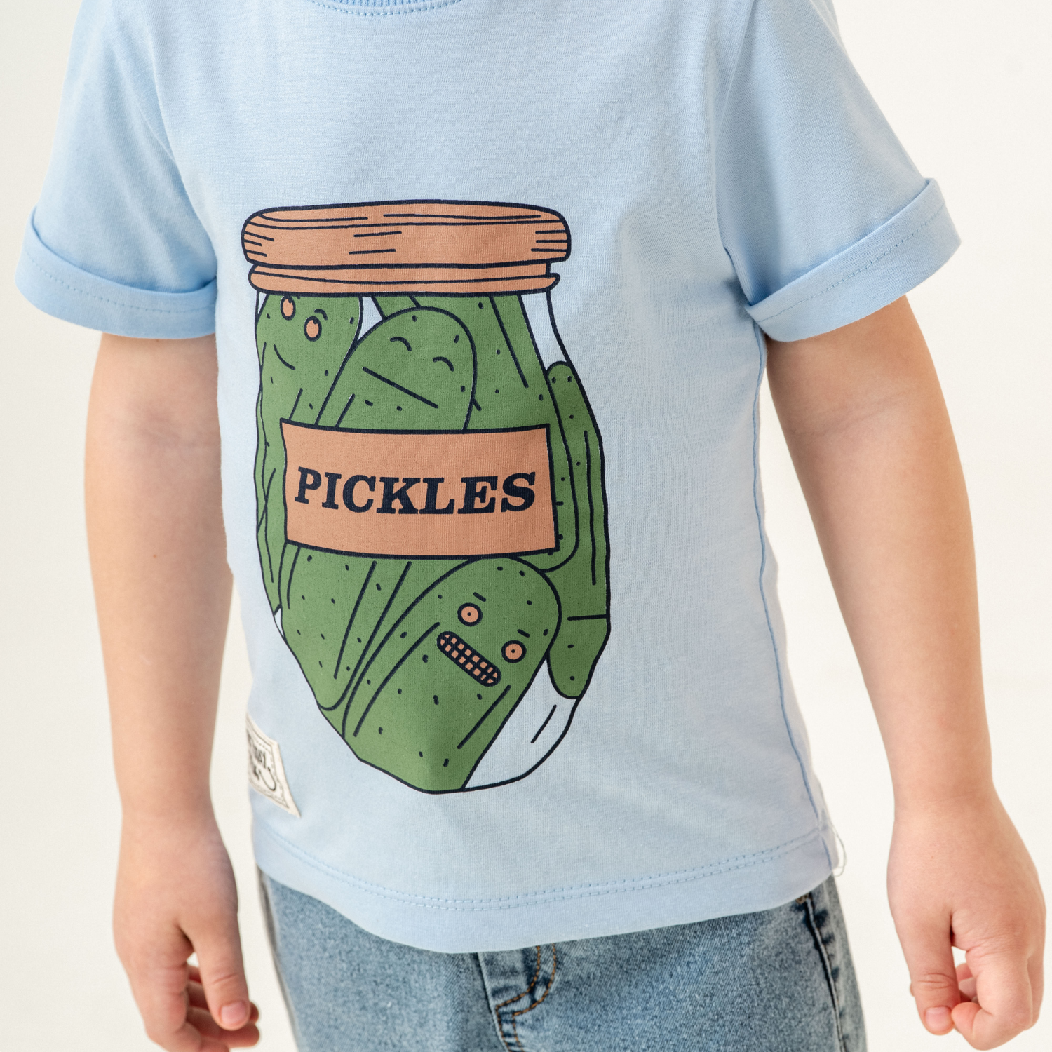 Toddler wearing a light blue T-shirt featuring a fun print of pickles with silly faces inside a labeled jar, paired with casual light denim jeans.