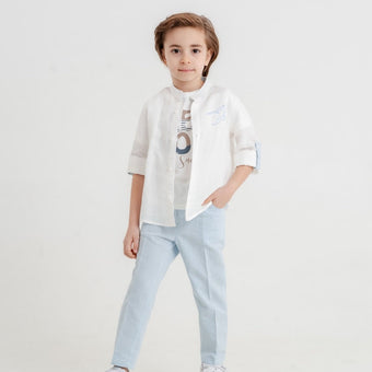 A child model dressed in a white button-up shirt with a bird embroidery over a printed tee, light blue pants & casual white shoes, hands in pockets.