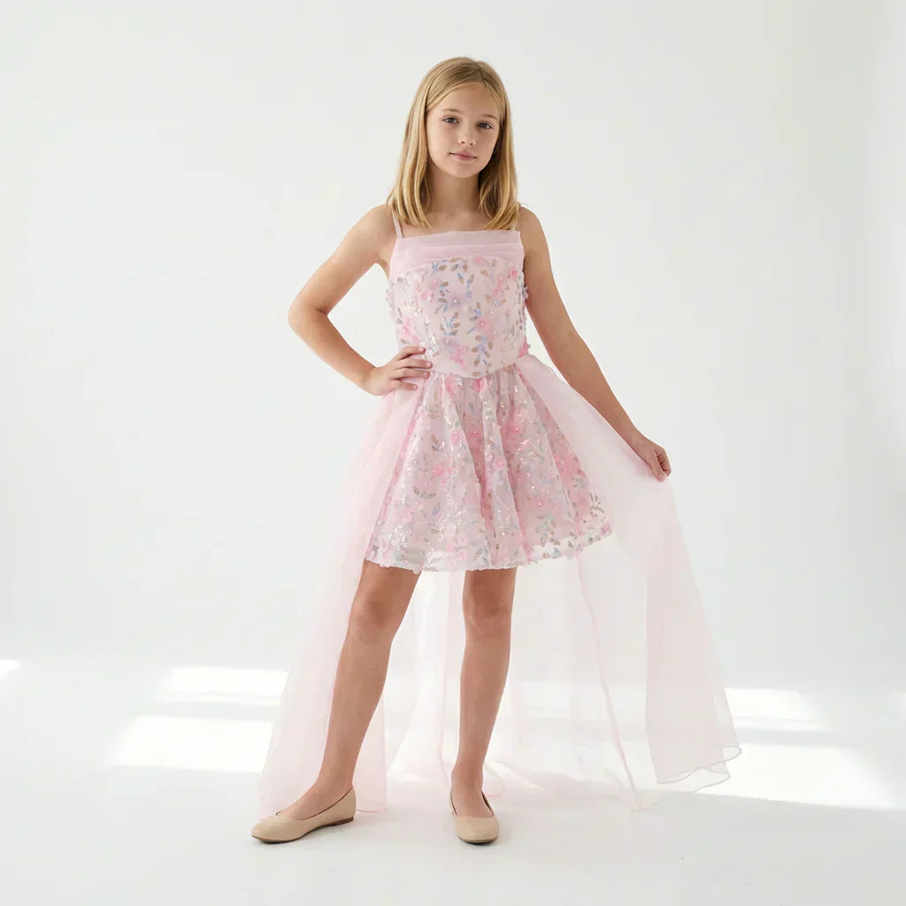Alt text: Girl in pink floral sequin mini dress with sheer tulle cape overlay and spaghetti straps, nude flats 