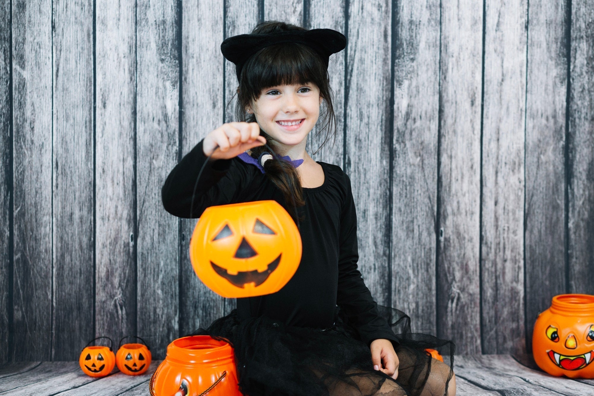 A smiling girl in a black cat Halloween costume holding a pumpkin bucket, sitting with festive decorations on a wooden backdrop.