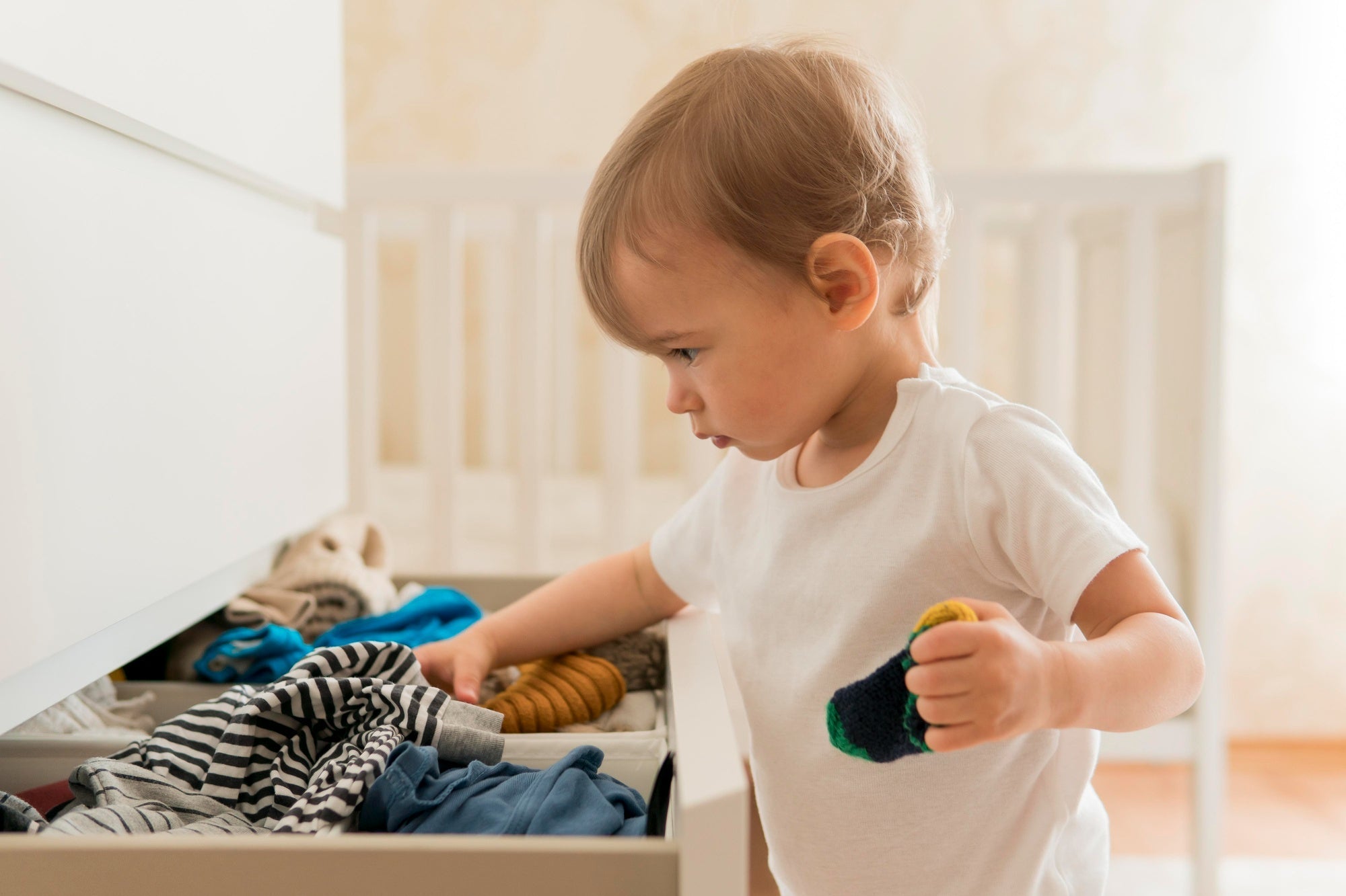 Toddler wearing a white T-shirt reaches into a dresser drawer filled with folded clothes in a softly lit nursery.