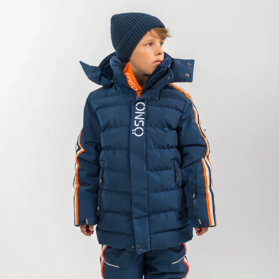Boy wearing navy ÖSNO ski jacket with orange stripe detail and knit beanie.