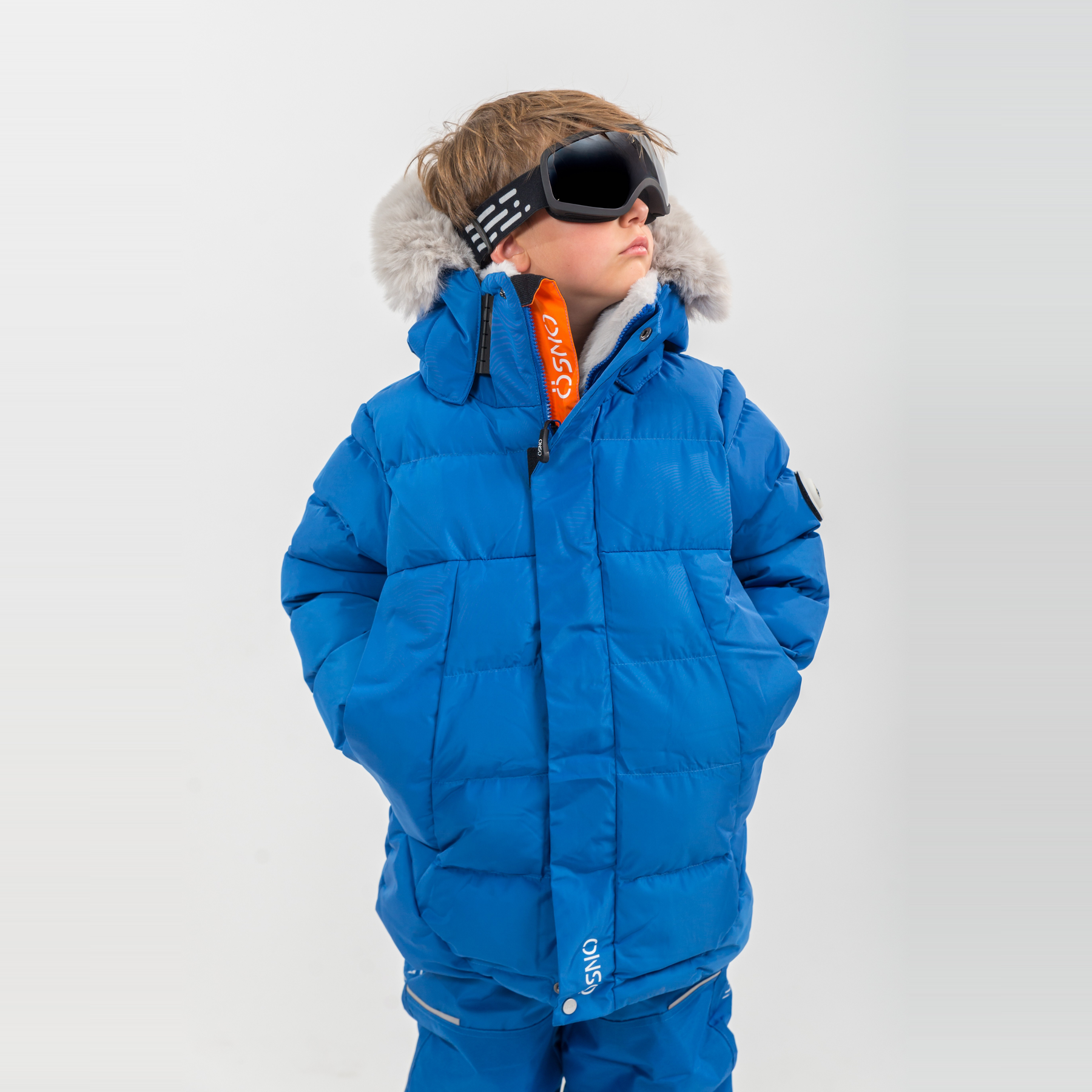 A boy in a bright blue toddler snowsuit with fur-trimmed hood and ski goggles, standing hands in pockets against a plain background.