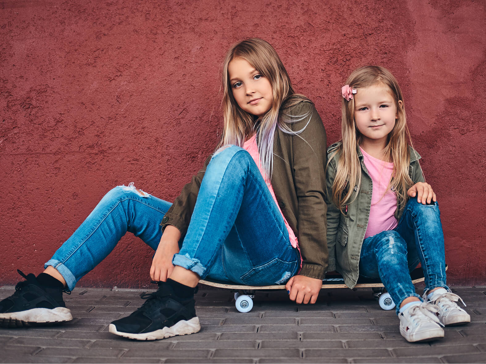 Two young girls with long hair wearing jeans and casual jackets sit together on a skateboard against a textured red wall outdoors.