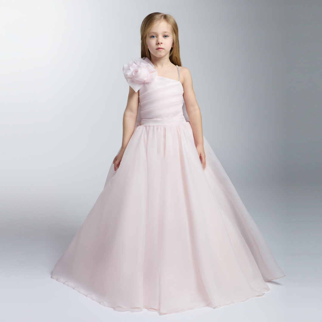 Young girl in a pale pink one-shoulder gown with a large floral accent and a full floor-length skirt.