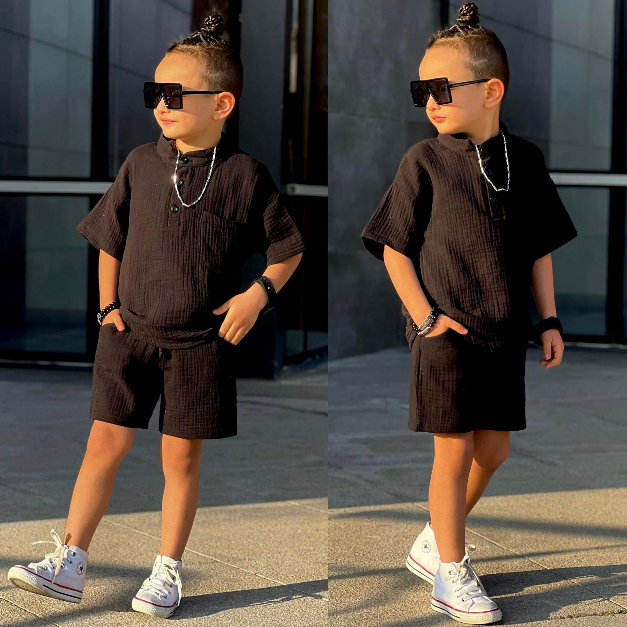 Stylish young boy wearing a black textured short-sleeve shirt & matching shorts, accessorized with oversized sunglasses, jewelry, and white sneakers.