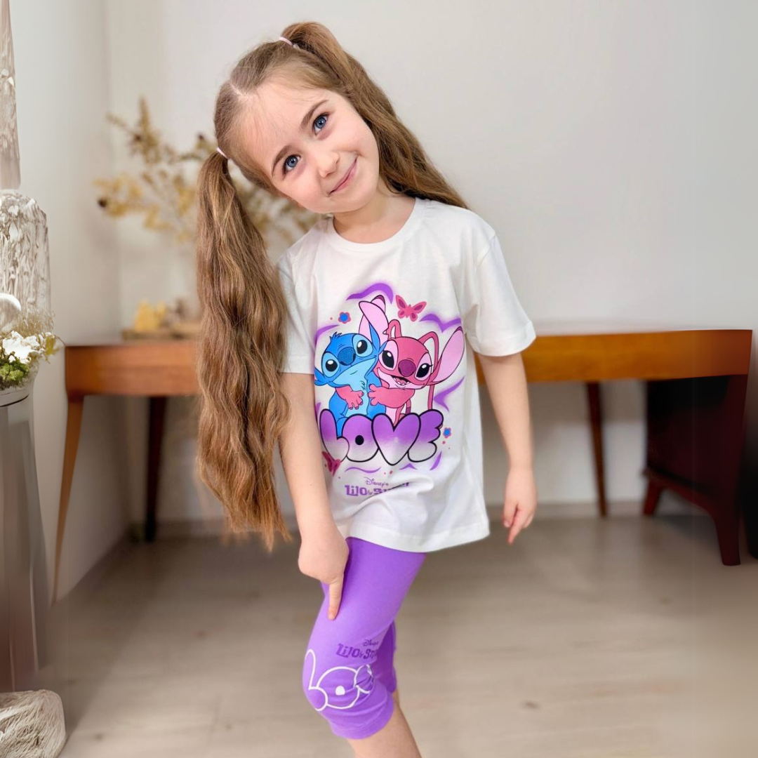 Young girl wearing a white cartoon graphic T-shirt and purple leggings, smiling indoors with long pigtails.