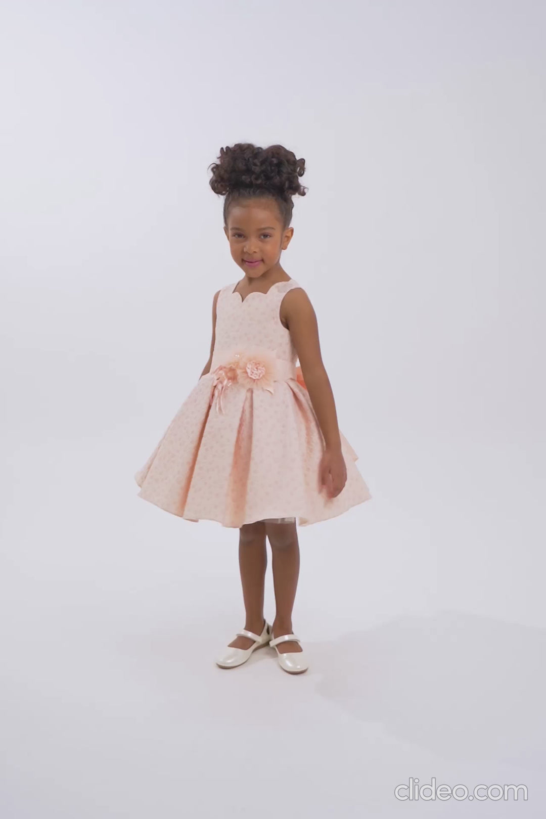 Young girl wearing a peach-pink sleeveless dress with a ribbon belt and flared skirt, standing gracefully in a white studio for a fashion photoshoot.