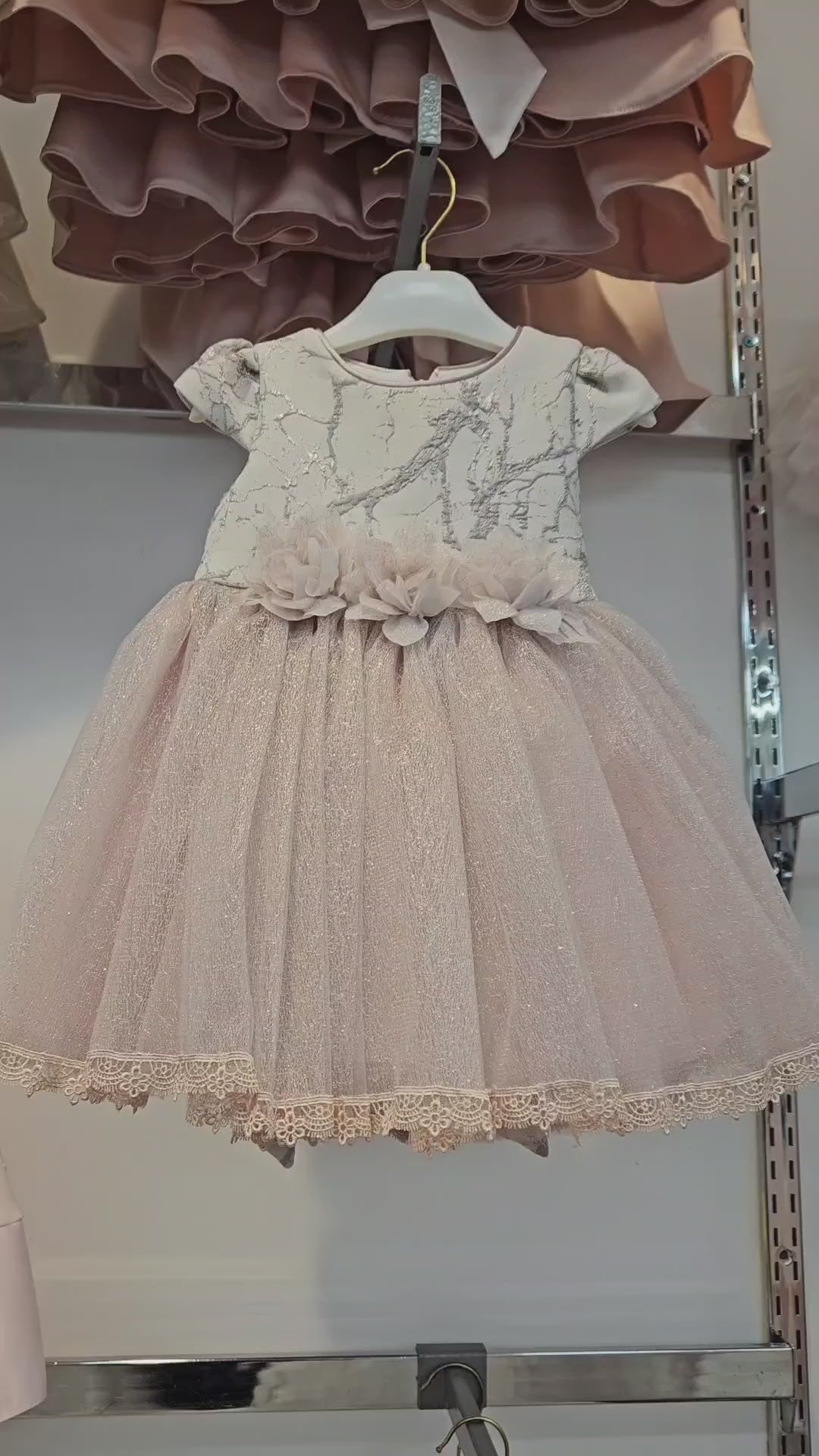 Elegant baby pink short-sleeve dress with lace bodice, ribbon waist, and tulle skirt, displayed on a hanger in a boutique children’s store.