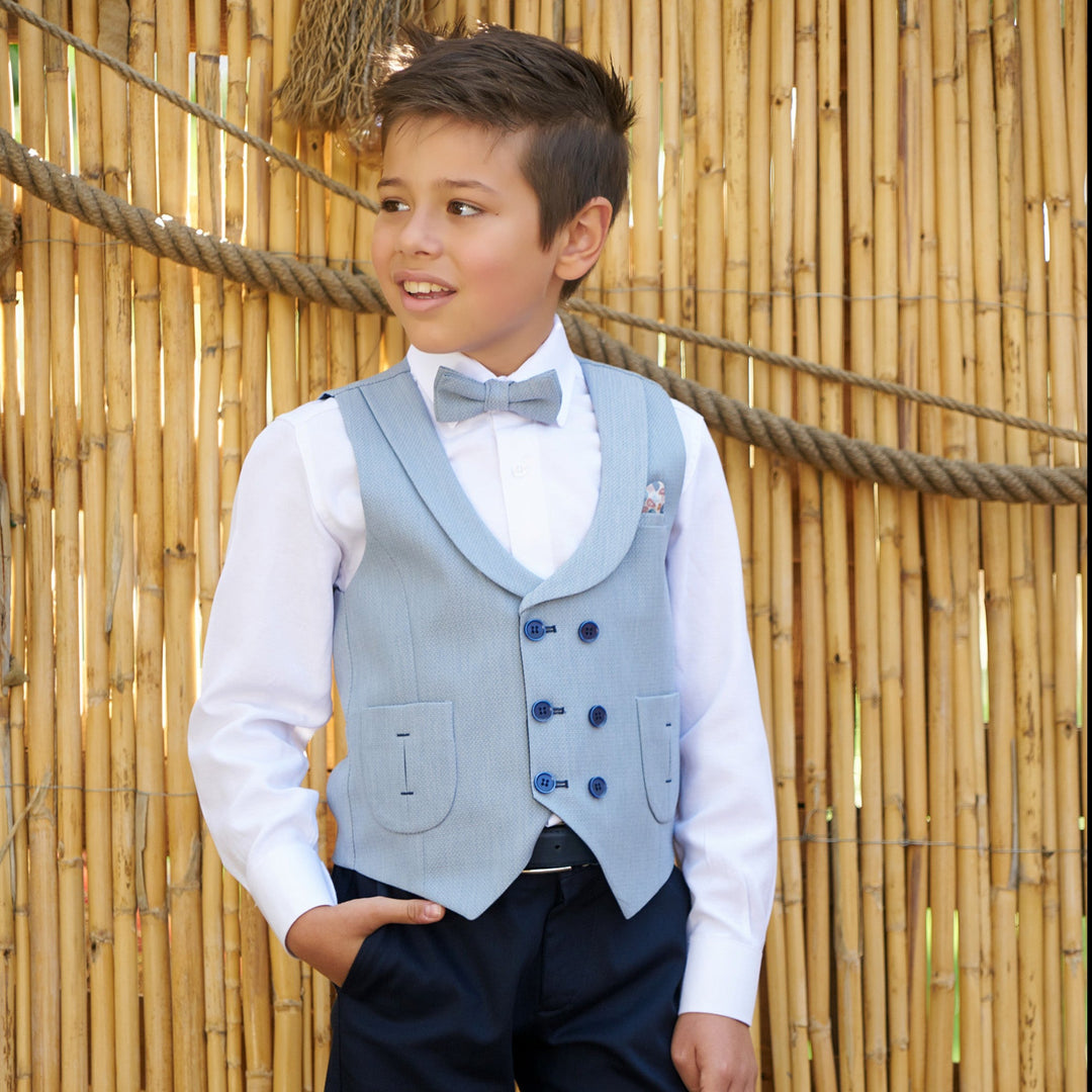 Young boy wearing a light blue vest, white shirt and bow tie, standing by a bamboo fence in a smart formal outfit.